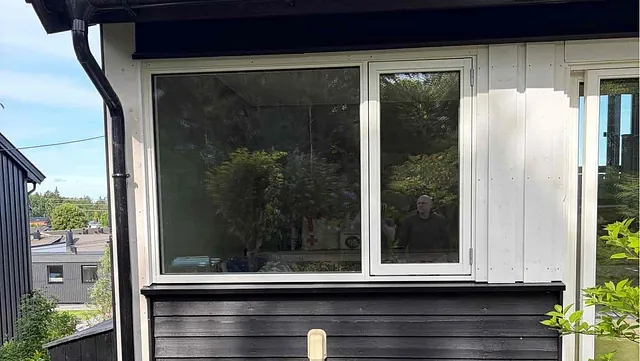 Hjørnet av et hus med vinduer, svart panel, rød takstein og frodig hage.