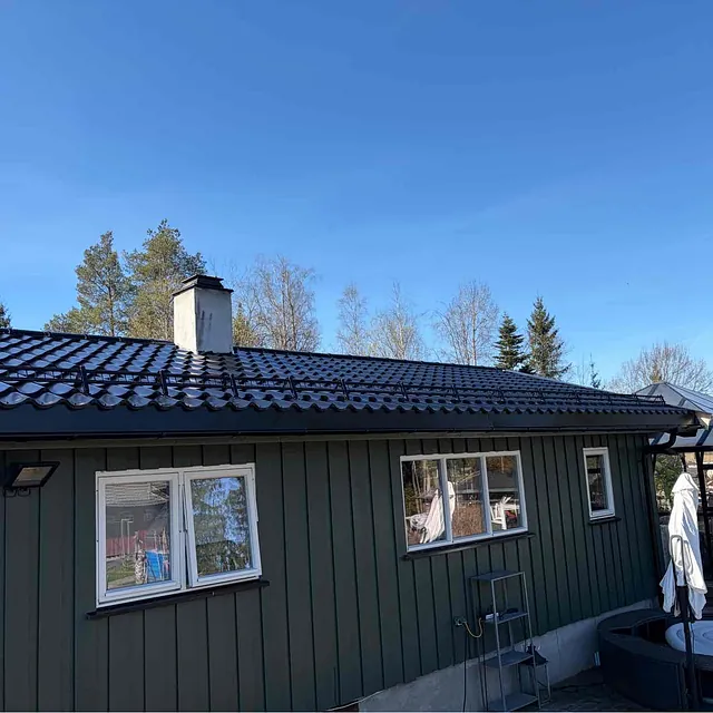 Tak på en grønns hus med blå himmel, trær i bakgrunnen og solfylte omgivelser.