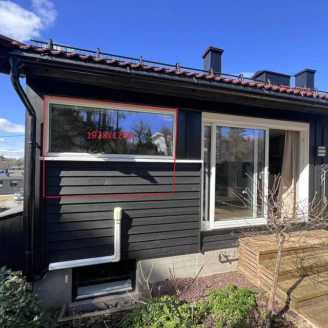Svart husvegg med vindu, terrasse og blå himmel i bakgrunnen.