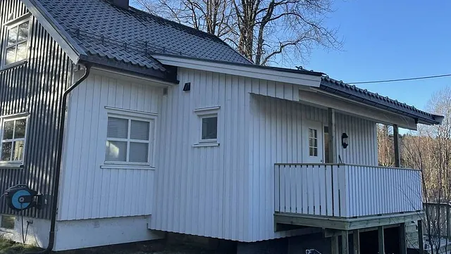 Hvit og svart bygning med balkong, omringet av trær under klar himmel. Byggematerialer foran.