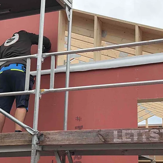 Arbeider på stillas med veggpanel under montering i nybygg. Delvis skyet himmel i bakgrunnen.