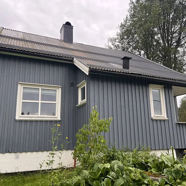 Blått hus med skråtak, stor hage og grønne planter, omkranset av trær.