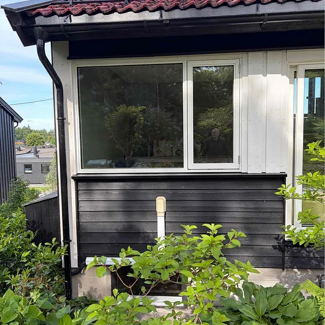 Hjørnet av et hus med vinduer, svart panel, rød takstein og frodig hage.