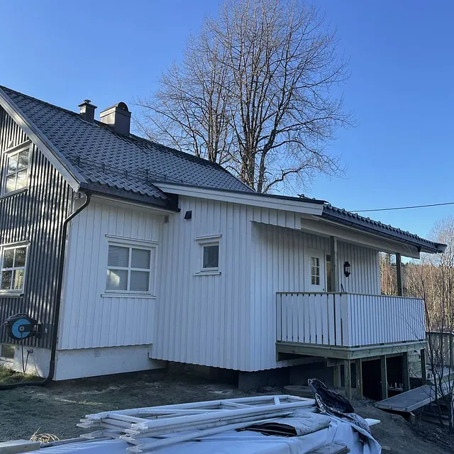 Hvit og svart bygning med balkong, omringet av trær under klar himmel. Byggematerialer foran.