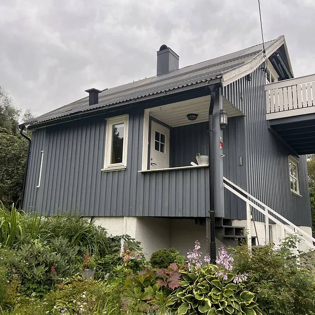 Grått hus med trapp og balkong, omringet av frodig hage og skyete himmel.