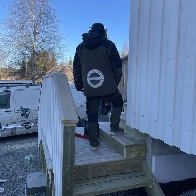 Person på trappen til en bygning, iført jakke og hørselsvern, med klar blå himmel bak.
